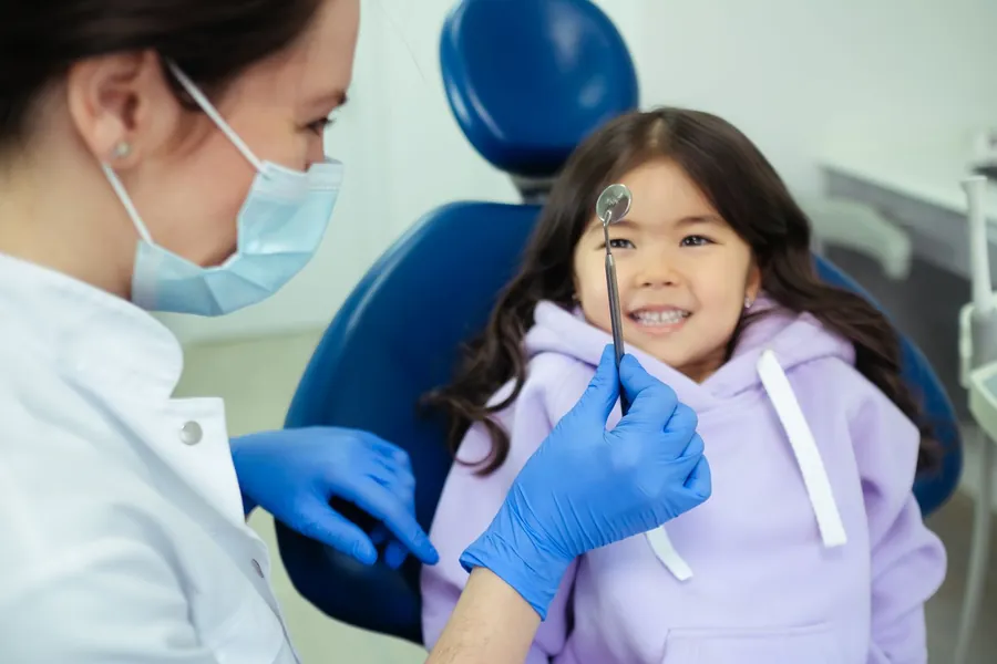 A little girl in a lilac hoodie smiling in the dental chair while the dentist holds up a mirror