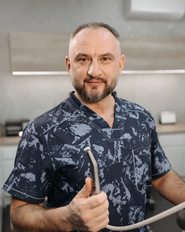 Greg Hollis in patterned scrubs, holding a dental tool and smiling at the camera