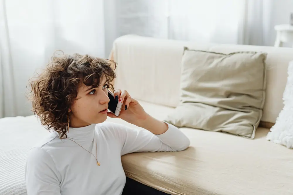 A woman listening on a phone call at home, seated on the floor beside a sofa in soft daylight
