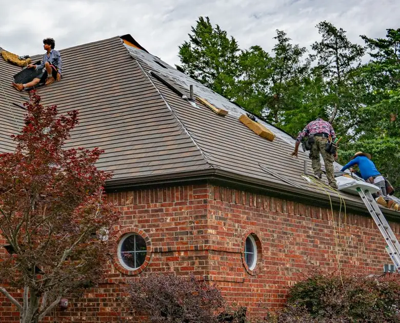 Roofing crew working on a brown shingle roof over a red brick house, ladders up
