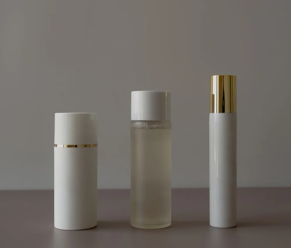 Three unmarked white and gold skin care bottles on a taupe shelf
