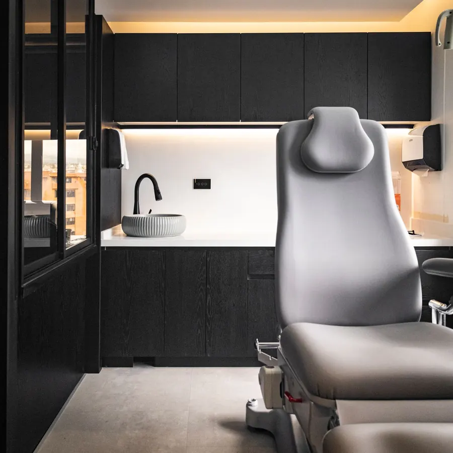 A grey reclining treatment chair beside a stone basin and black cabinets lit from below