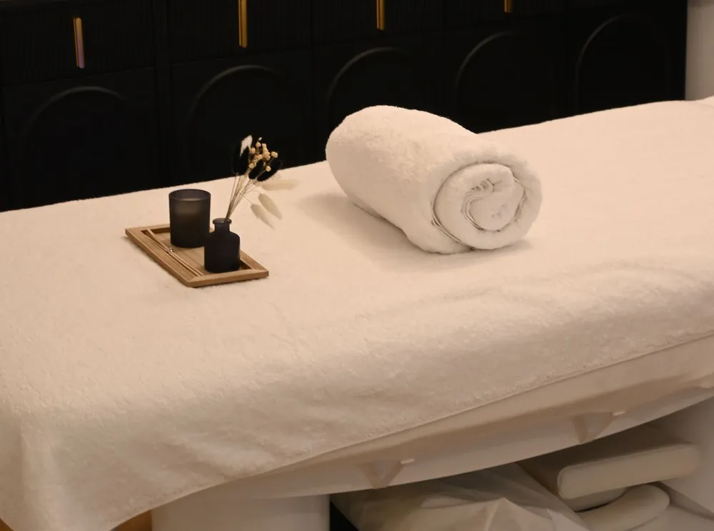 A treatment bed with a rolled white towel, a small tray and dried grasses in front of a black cabinet with brass handles