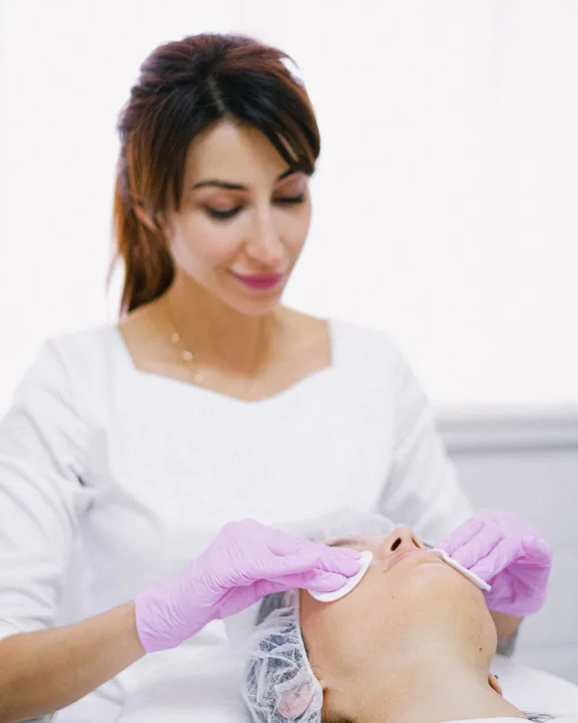 Leila Farah, licensed aesthetician, in a white tunic and pink gloves cleansing a client’s face