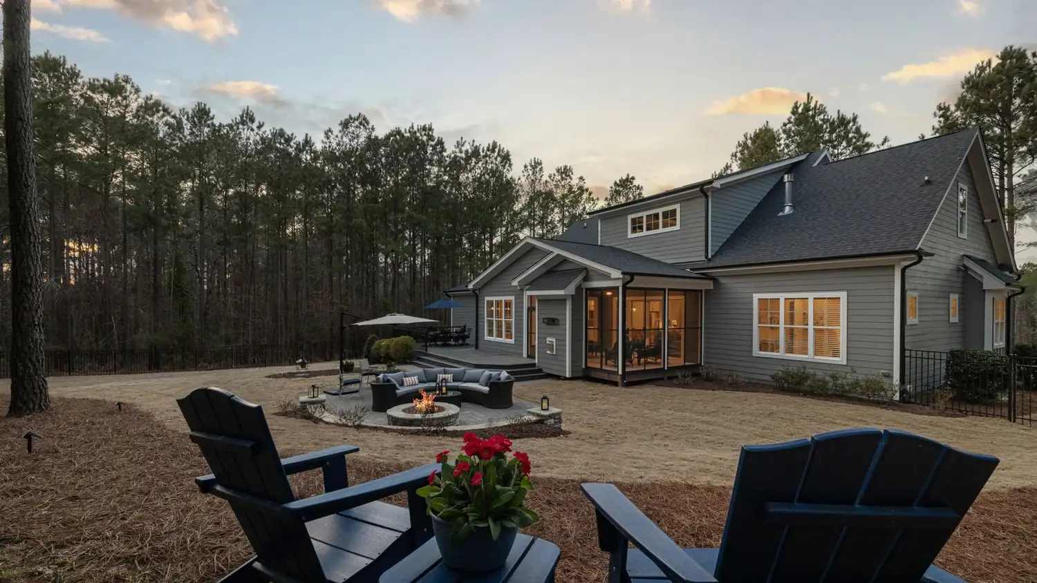A stone fire court with curved seating at dusk behind a gray home, ringed by planted beds and tall pines