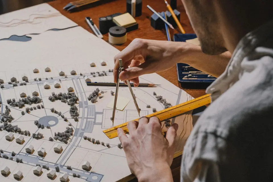 A designer drawing on a scale site plan with a compass and ruler, small model trees and buildings on the board